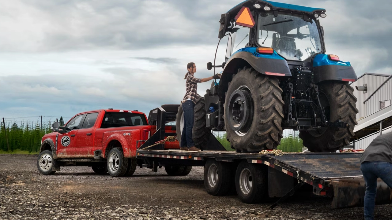 Ford F-250 rouge avec remorque plateau transportant un tracteur agricole bleu, avec plusieurs travailleurs assistant au déchargement.